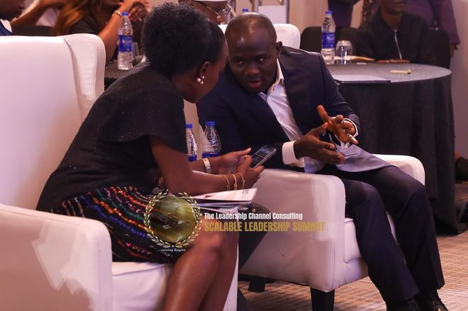 Man and woman in discussion, seated in white chairs.  Man gestures, woman looks on. Setting is a conference.