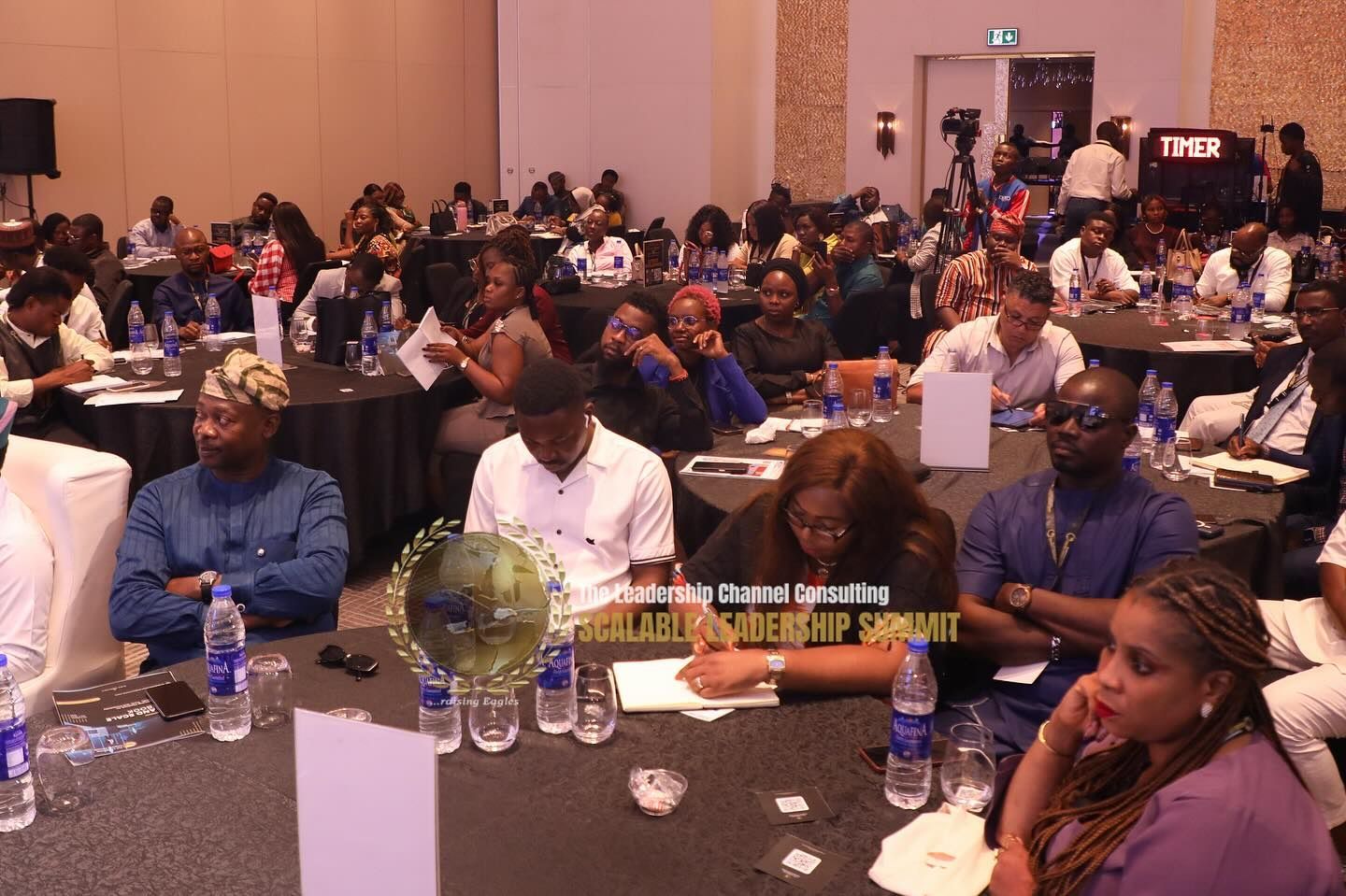 Conference attendees seated at tables, taking notes. Brightly lit room.