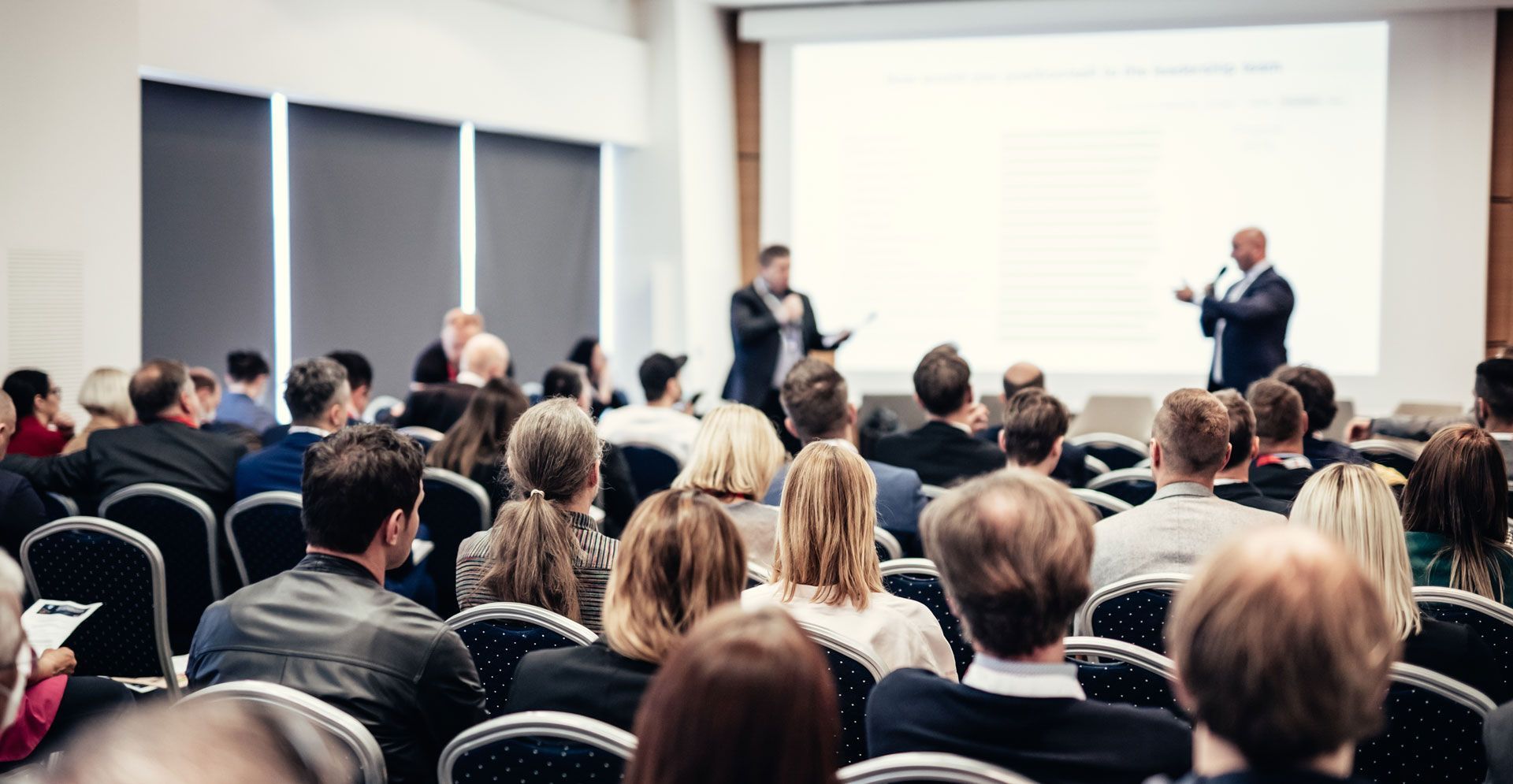 Audience at a conference listens to two speakers presenting, projection screen visible, indoors.