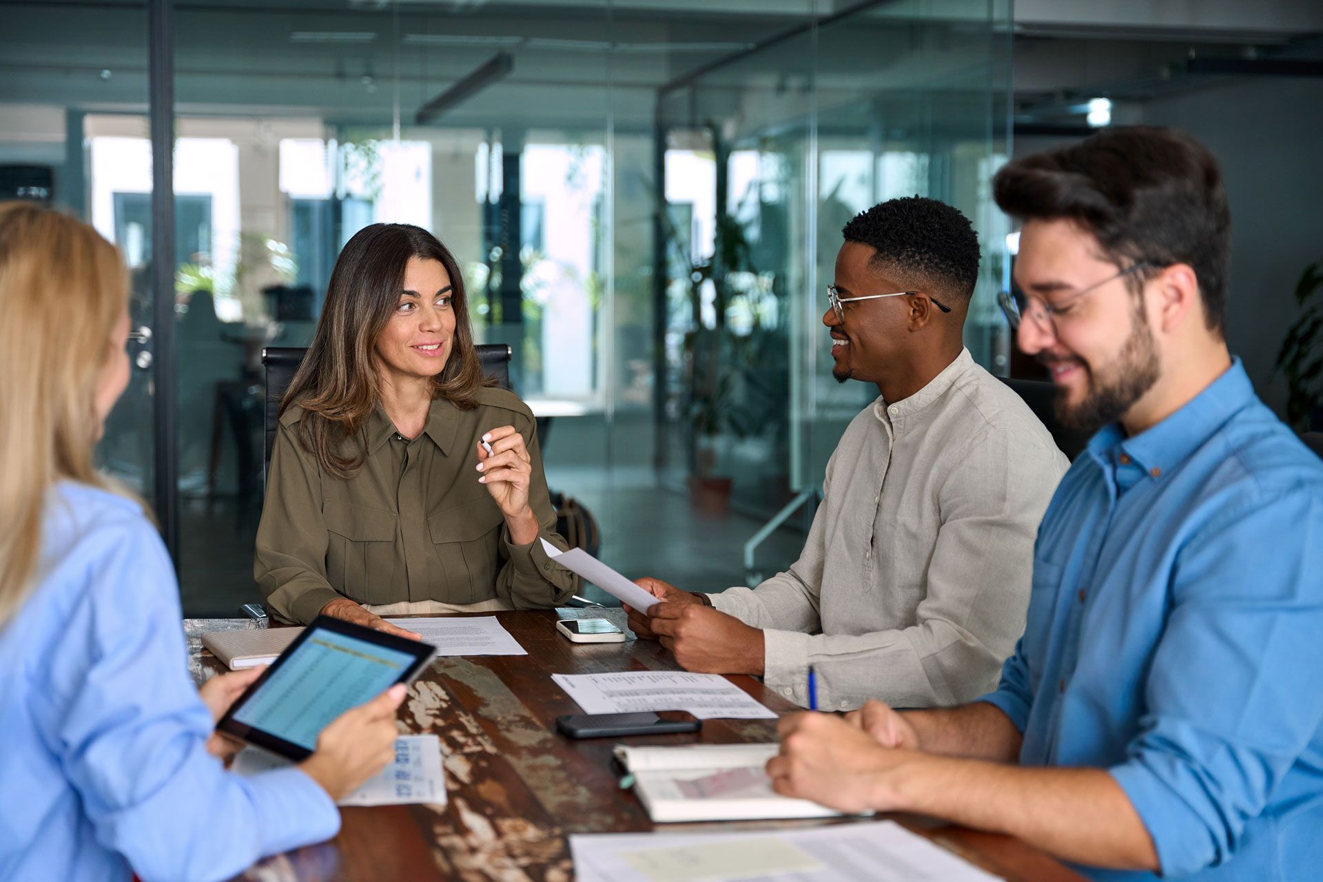 Four diverse colleagues in a meeting at a table, discussing documents and using a tablet.