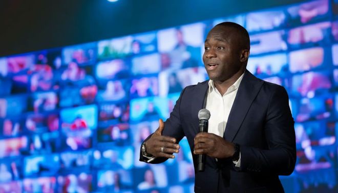 Man in suit speaks into a microphone with a virtual audience on a large screen behind him.