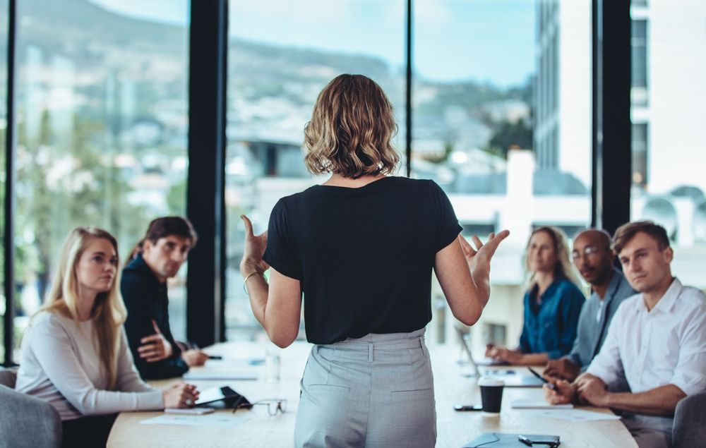 A woman is giving a presentation to a group of people sitting around a table.