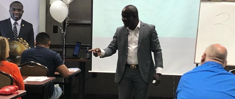 A Black man in a suit lectures at a class, pointing. People sit at desks, another photo is behind him.