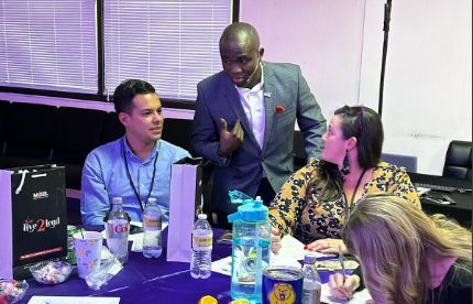 Three people at a table discussing something. One man stands gesturing while two others sit, looking at him.