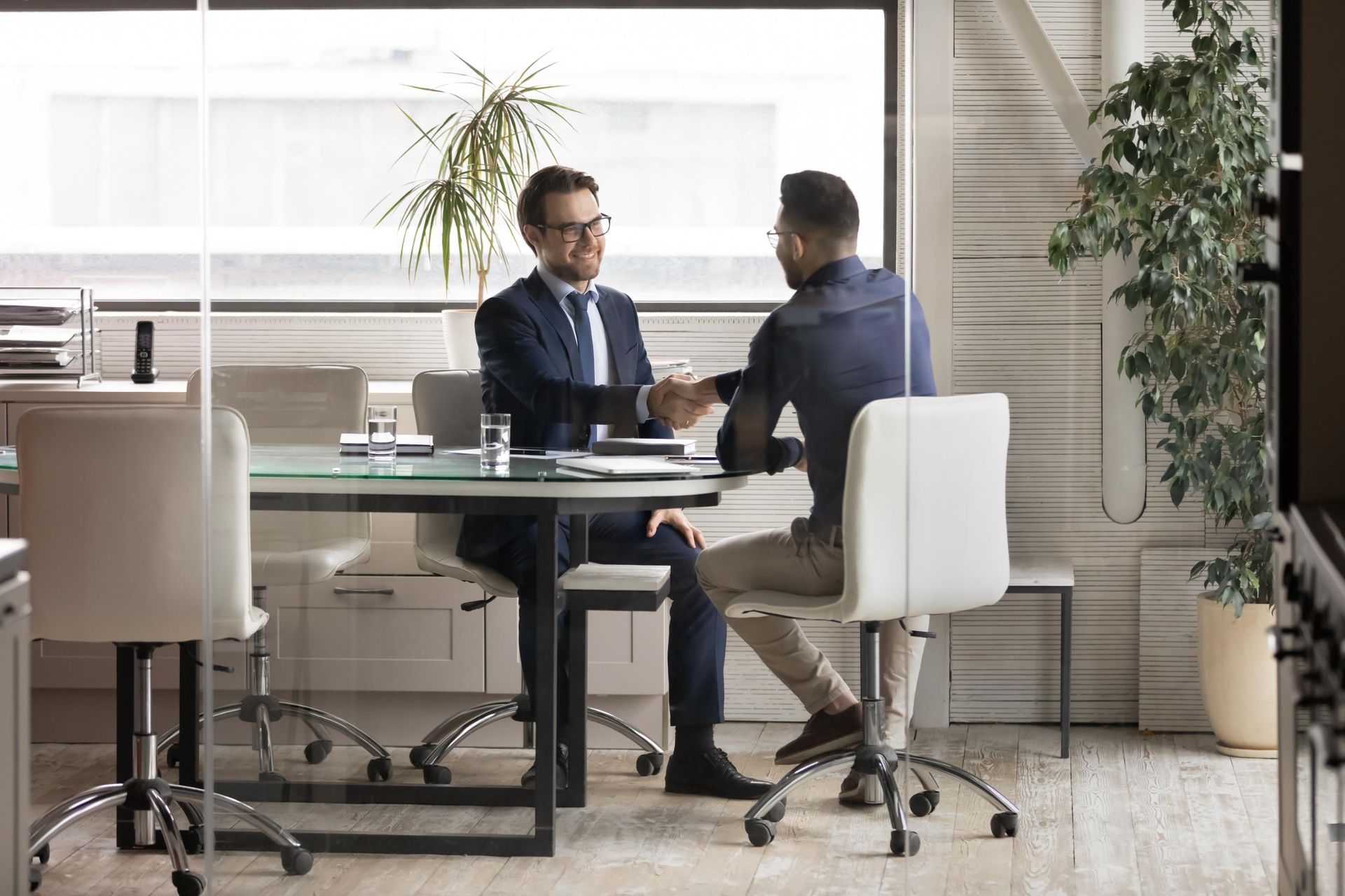 Two men are shaking hands while sitting at a table in an office.