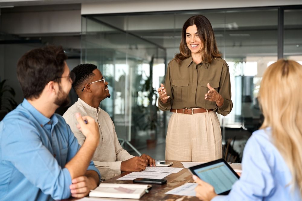 A woman is giving a presentation to a group of people sitting around a table.