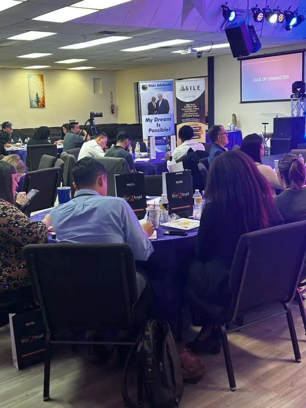 People seated at tables in a conference room, some looking towards a stage with a screen.