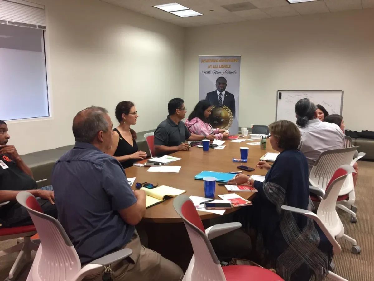 People seated around a conference table, engaged in discussion. A poster of a man is visible on the wall.