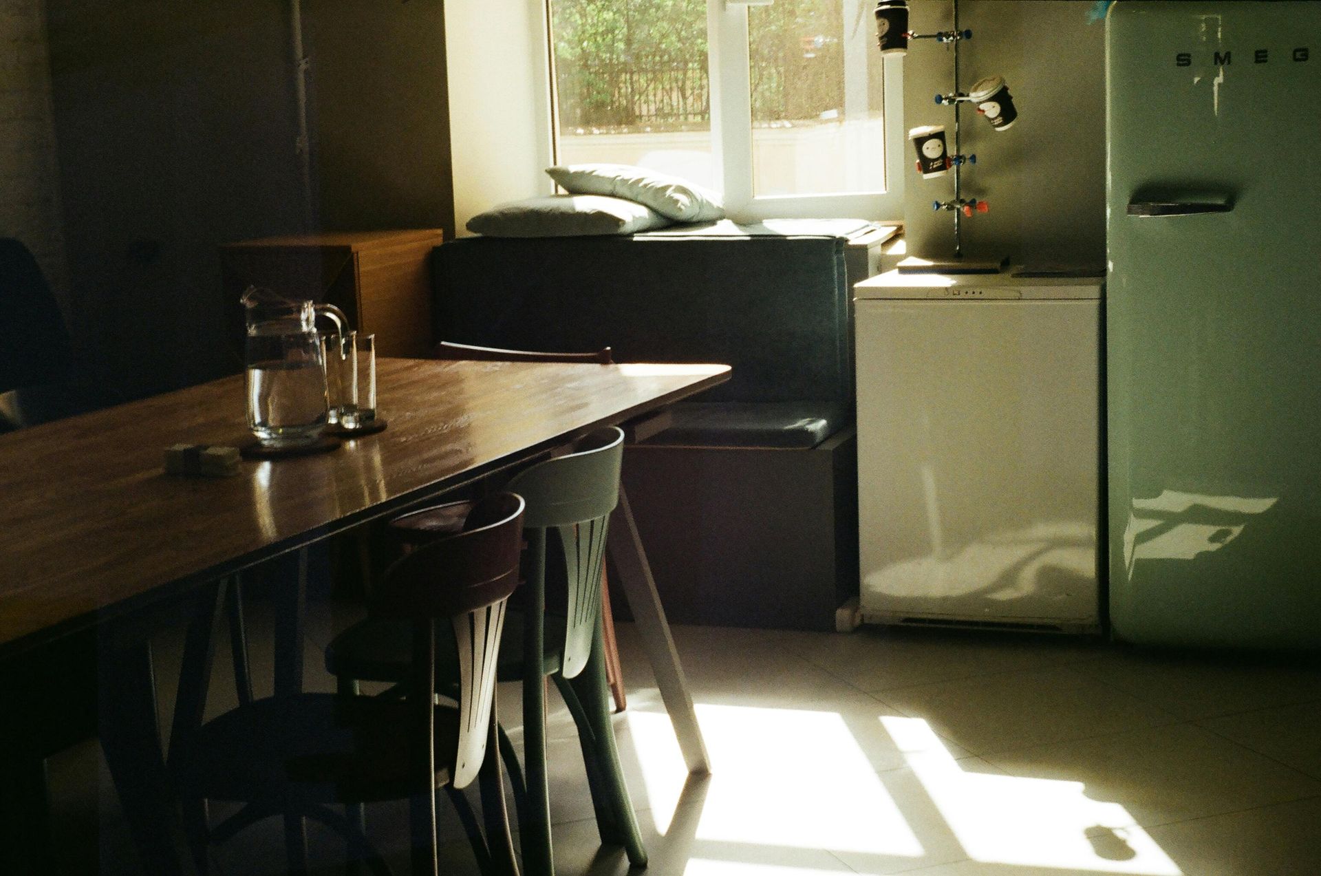 Un coin cuisine avec une table en bois, des chaises, une fenêtre, un réfrigérateur et la lumière du soleil qui entre.