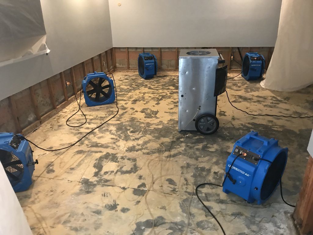 Several blue floor dryers and a dehumidifier in a room with a damaged floor, likely drying out after water damage.