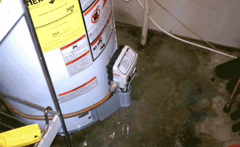Water pooling on a floor beside a large water heater and a small pump or valve assembly.
