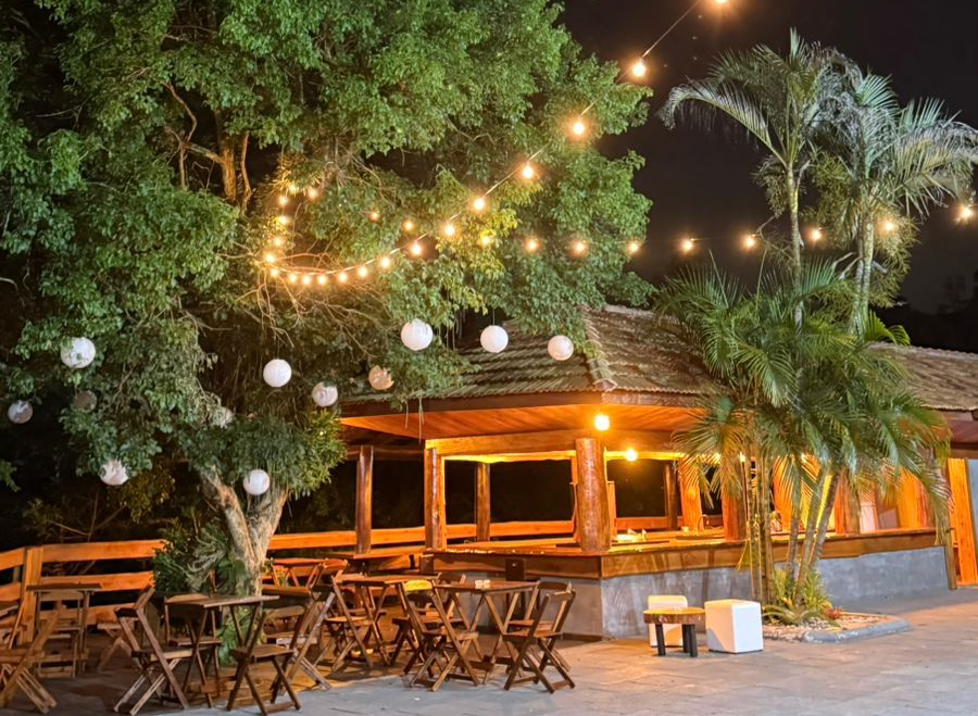 Restaurante ao ar livre à noite com luzes de corda, árvores e mesas de madeira.