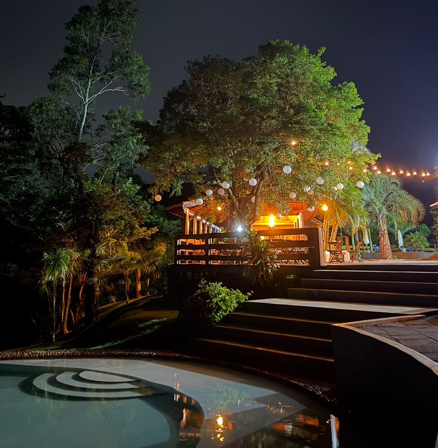Cena noturna ao ar livre: Prédio com luzes de corda perto de uma piscina, situado entre árvores, com escadas que levam ao andar superior.