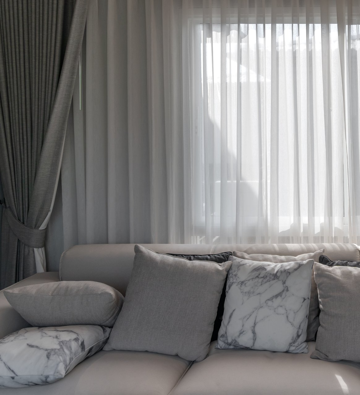 Grey couch with cushions in front of window covered with sheers curtains, framed by tied back gray block-out curtains.