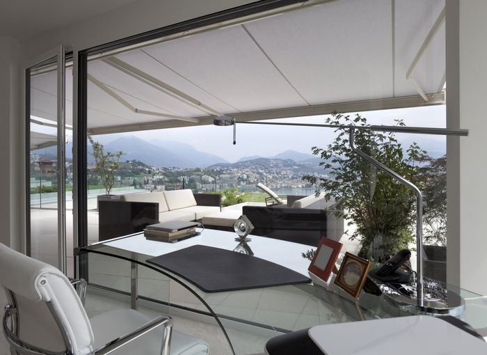 Doors of office with white chair and glass desk opening out onto verandah with awning covering outdoor lounge, looking out over view of town and mountains.