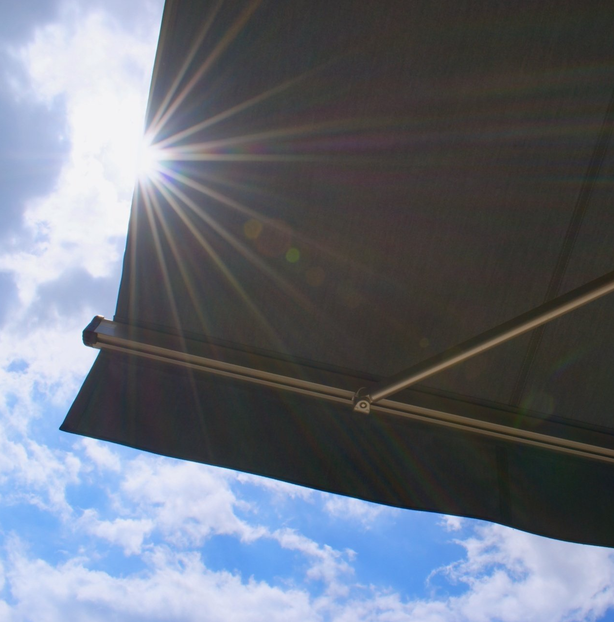 Upward view of black outdoor awning, with sun shining brightly in slightly cloudy blue sky.