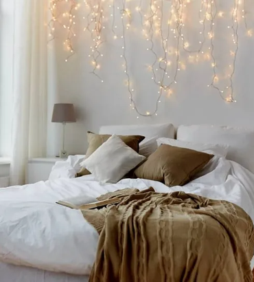Fairy lights shining above comfy bed with brown bedspread, framed by white curtain and lamp.