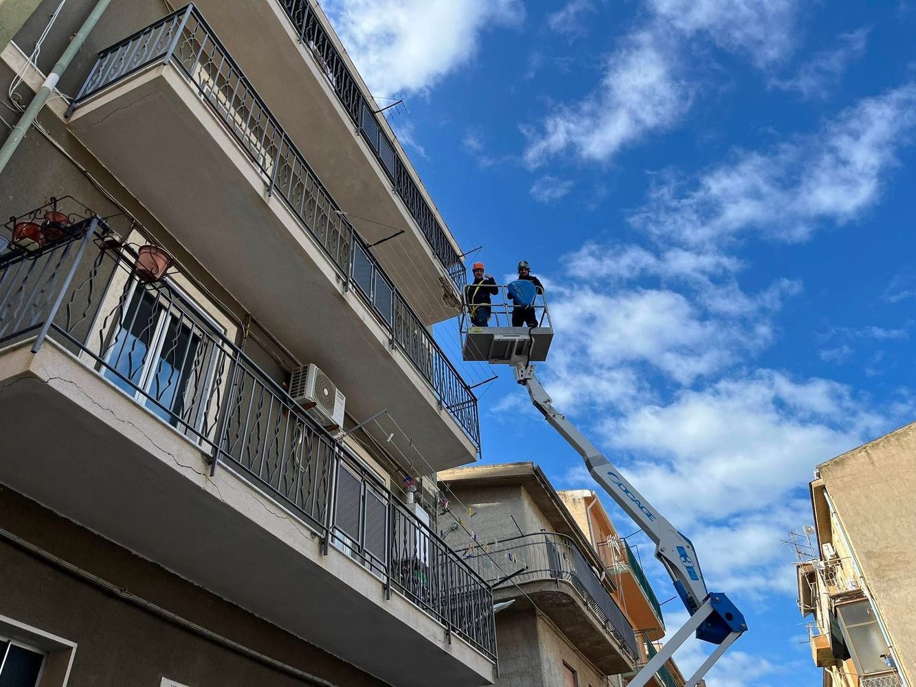 Due persone su un ascensore riparano la facciata di un edificio sotto un cielo azzurro e nuvoloso.