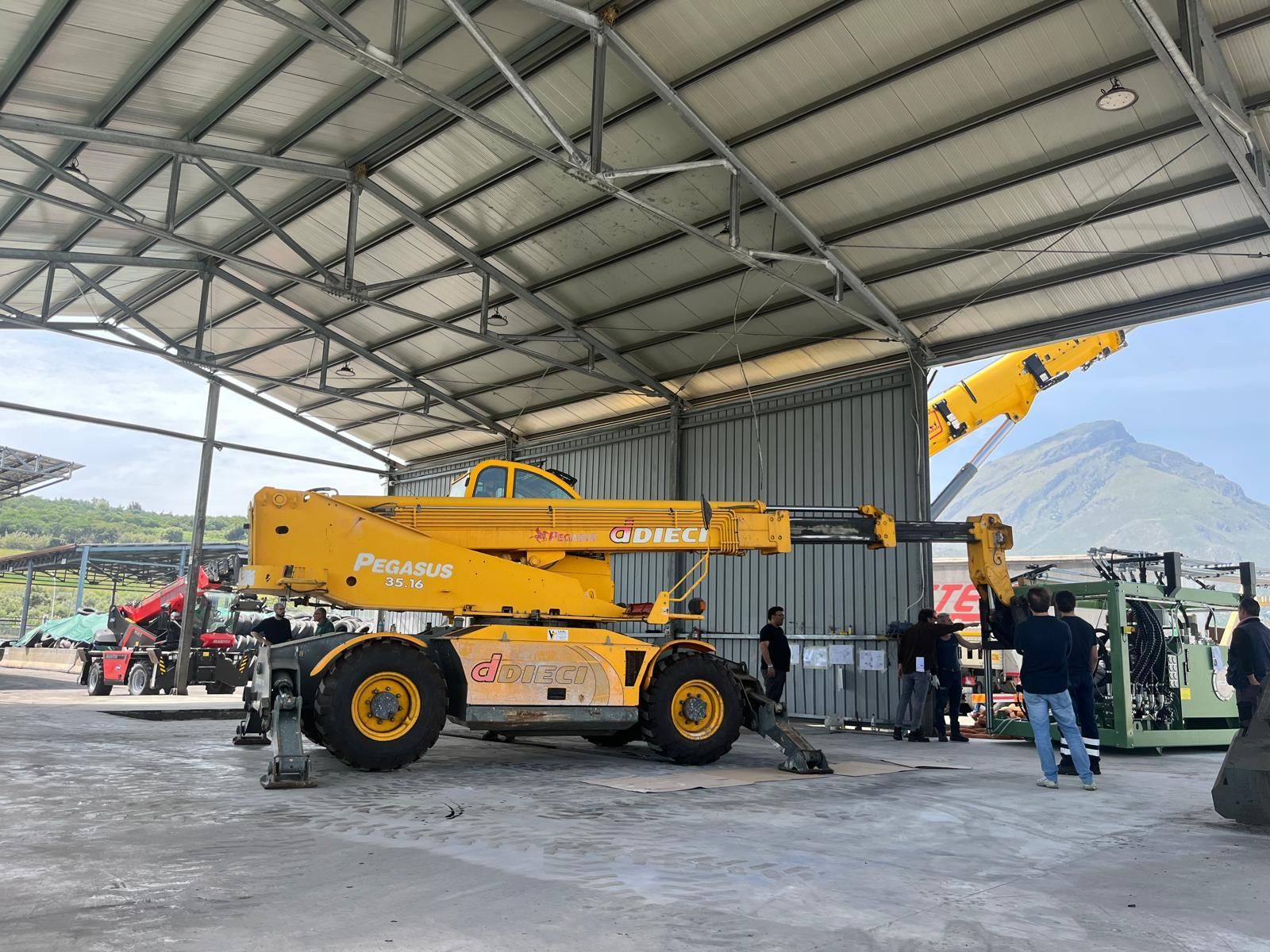 Due operai su una piattaforma elevatrice gialla, che installano qualcosa su una struttura metallica del tetto. All'aperto, soleggiato.