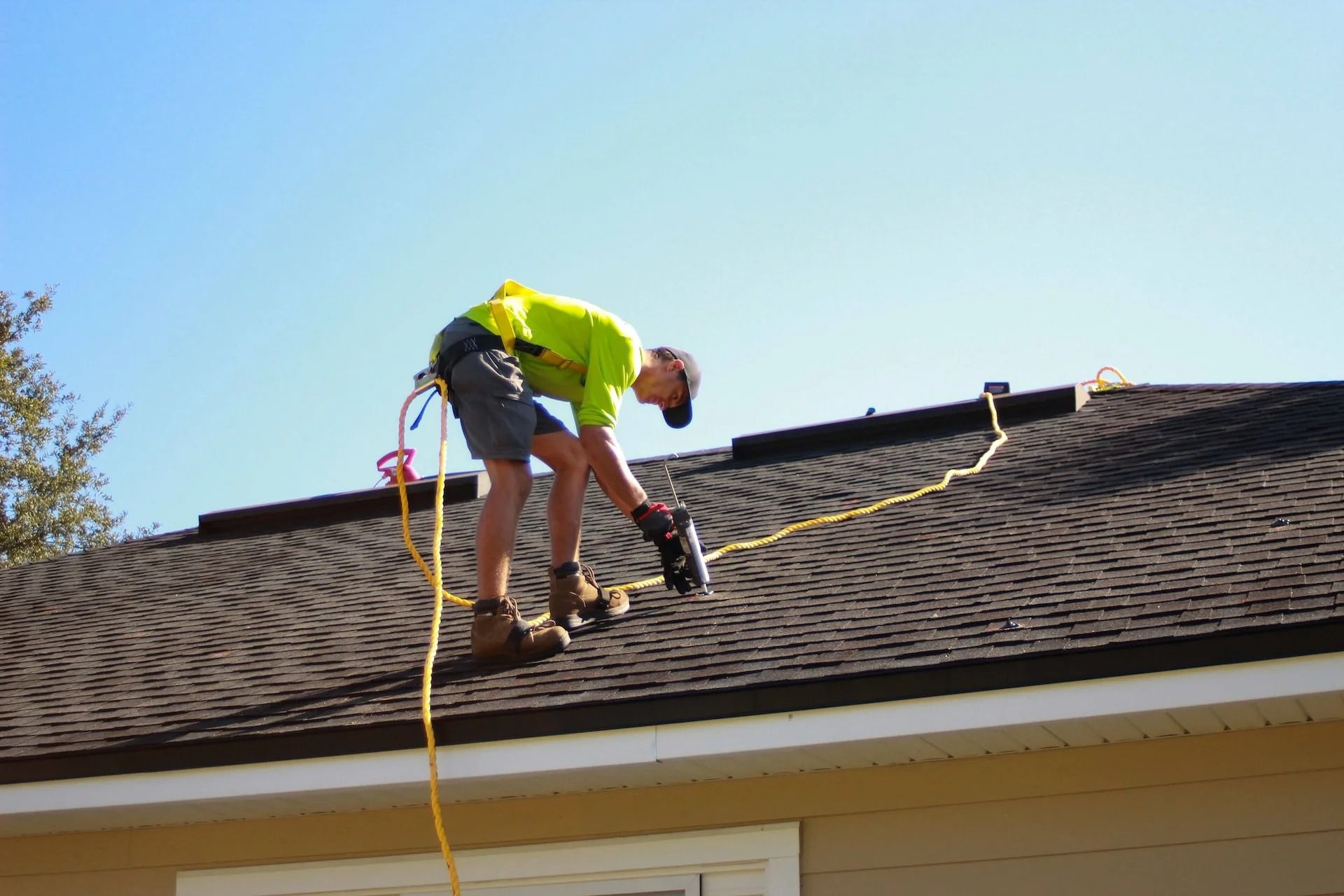 Worker on a shingled roof using a power tool with a safety harness and cord.