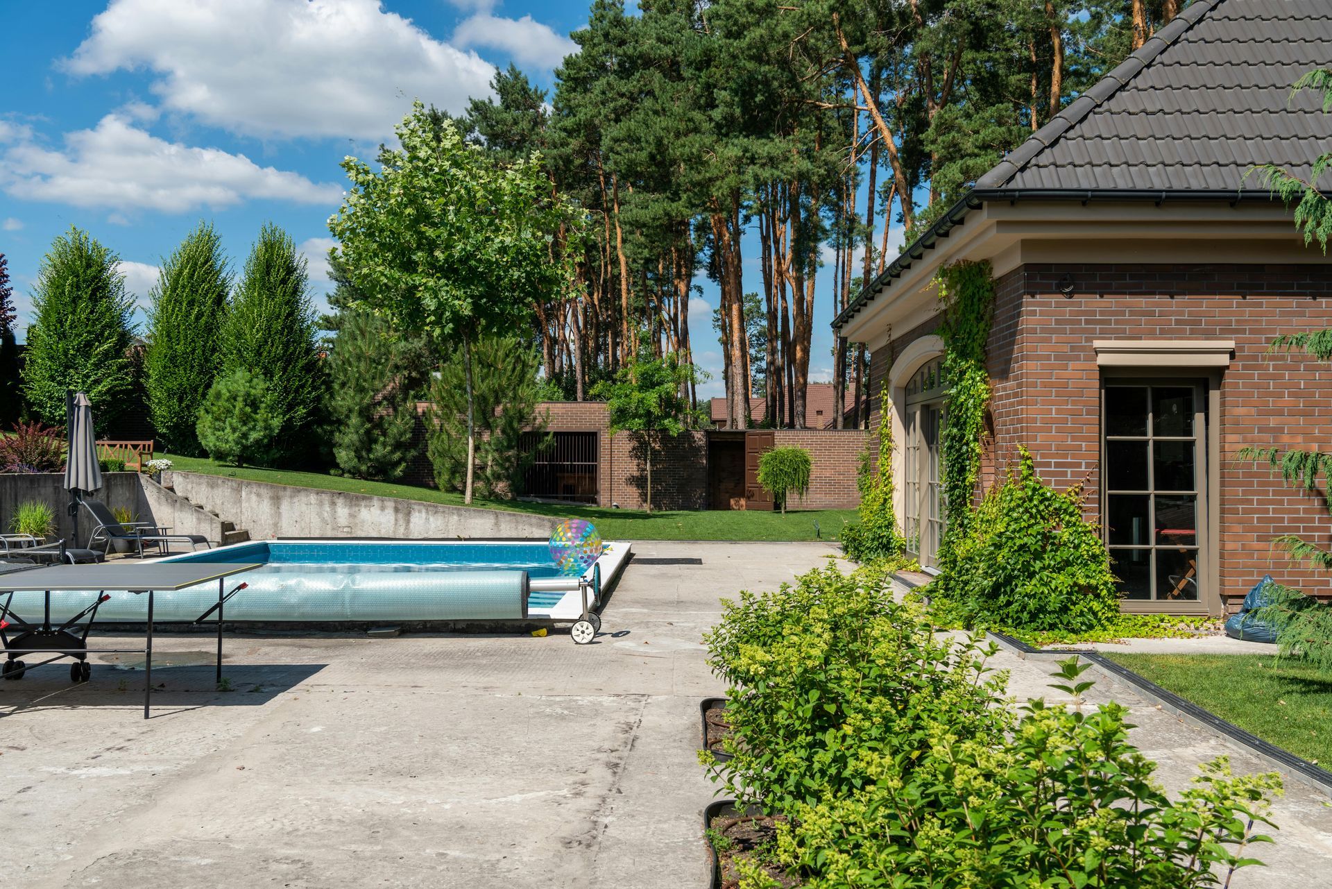 Backyard patio with a covered pool, green shrubs, and a brick house under a blue sky