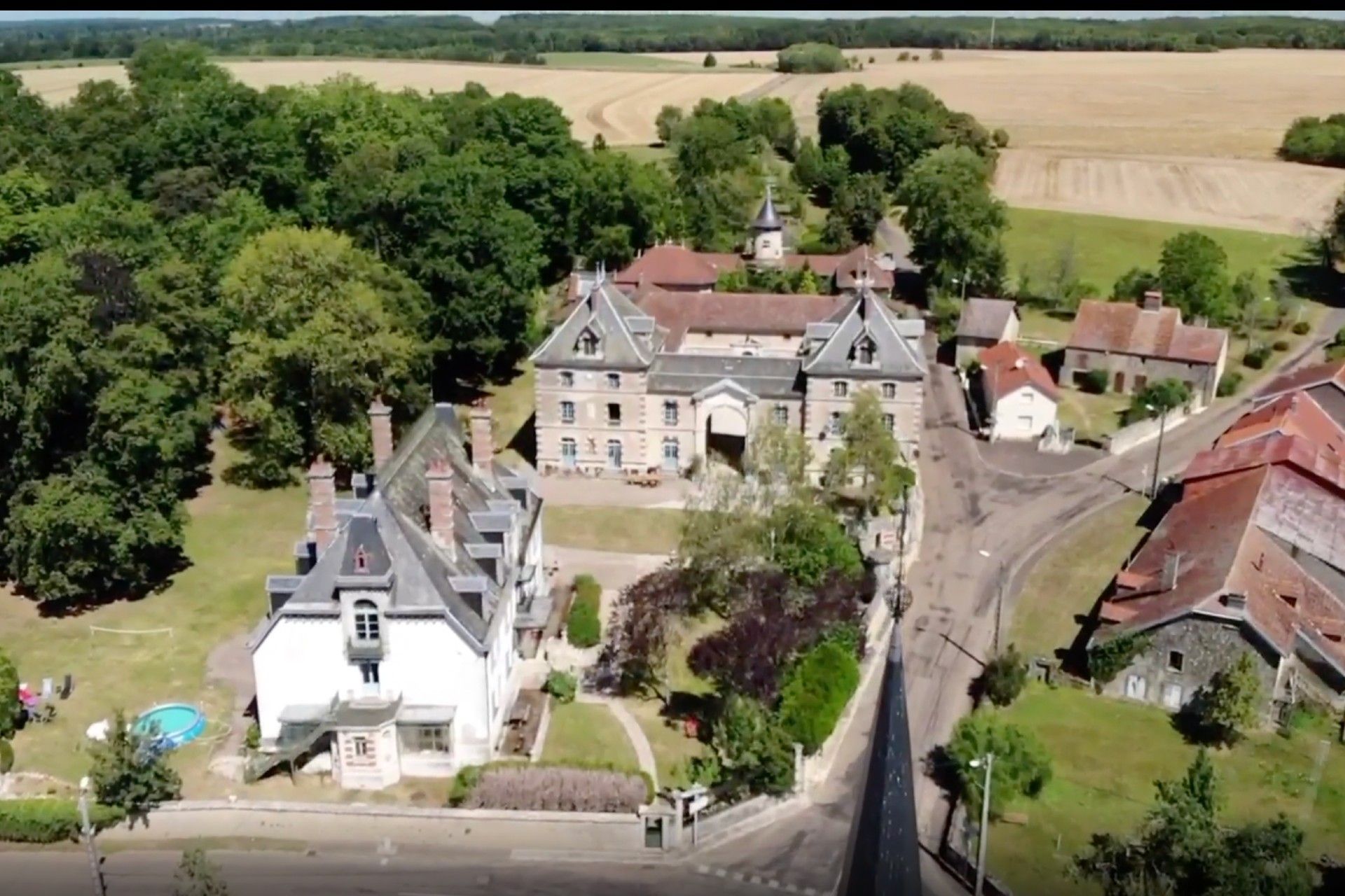 Groot wit huis met blauwe luiken en een donker dak op een groen gazon. Aan de linkerkant loopt een veranda.