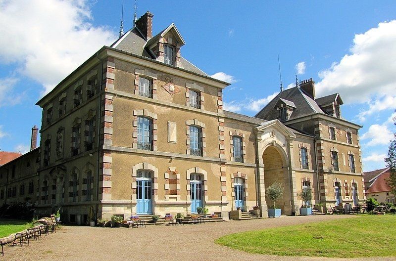 Groot wit huis met blauwe luiken en een donker dak op een groen gazon. Aan de linkerkant loopt een veranda.