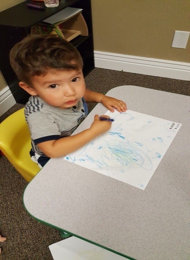 A young boy is sitting at a table drawing with a crayon.