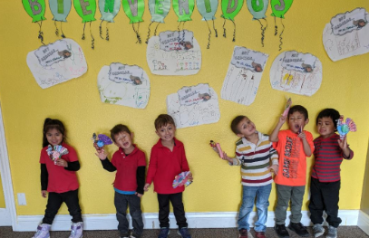 A group of children are standing in front of a yellow wall with balloons hanging from it.