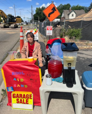 Girl at a lemonade stand and garage sale on a street with construction.