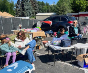 People at a yard sale, sitting and selling items in a parking lot on a sunny day.
