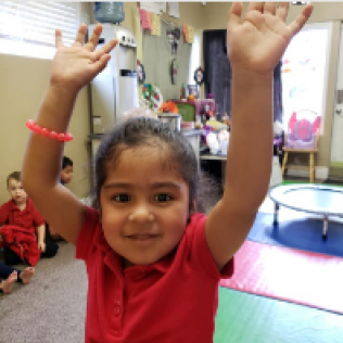 A Little Girl in a Red Shirt is Raising Her Hands in the Air