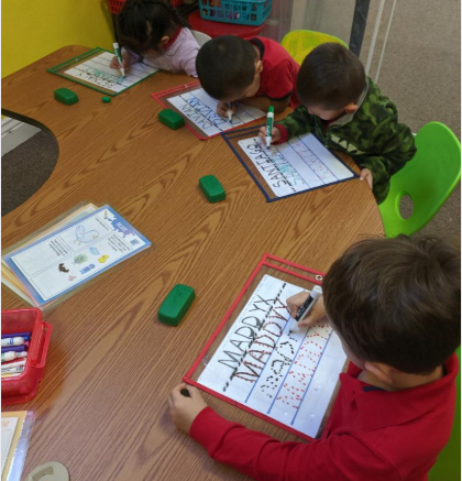 A group of children sit at a table writing on a piece of paper that says maddy