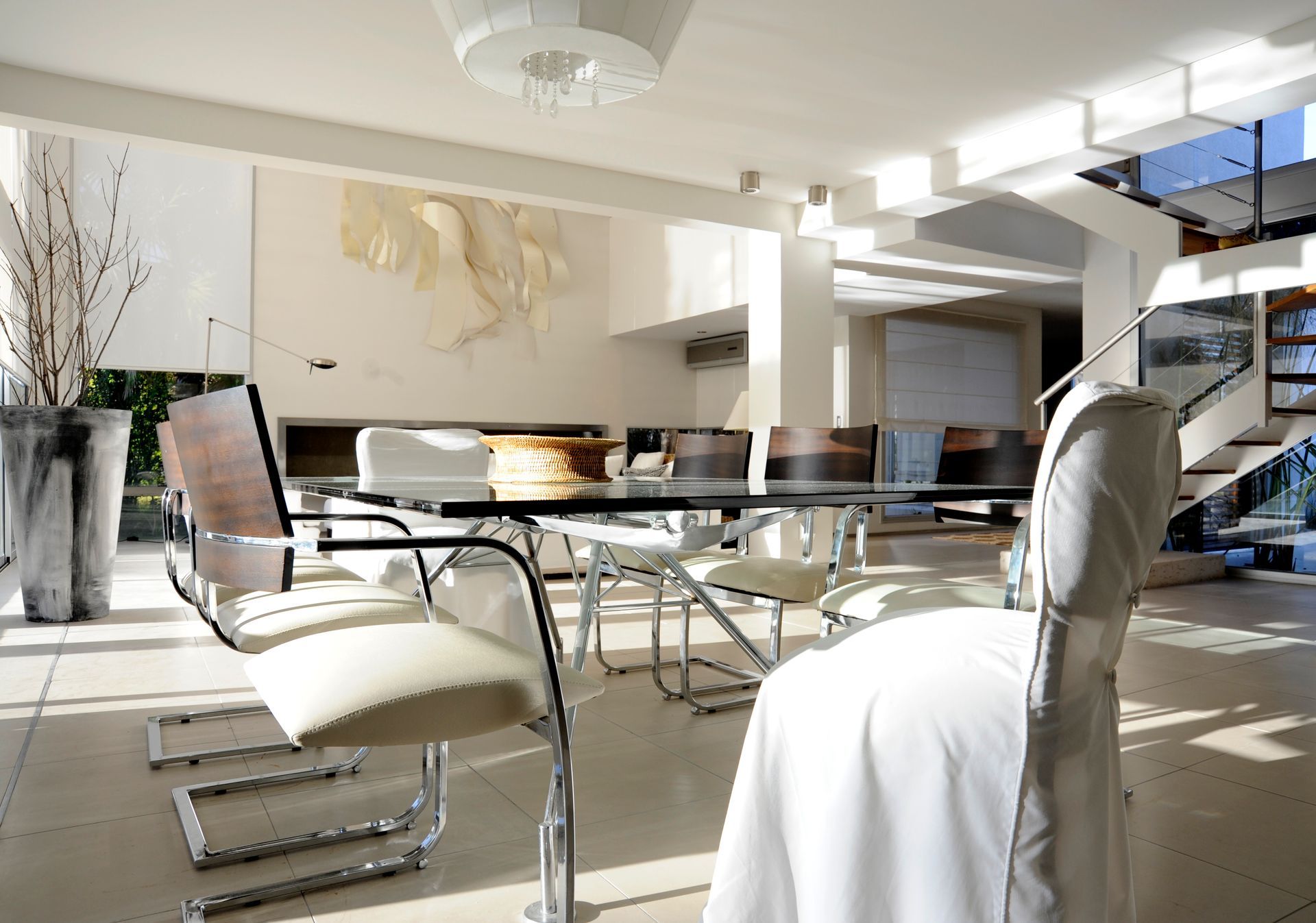 Comedor luminoso con mesa de cristal, sillas cromadas y obras de arte moderno. La luz del sol entra a raudales.