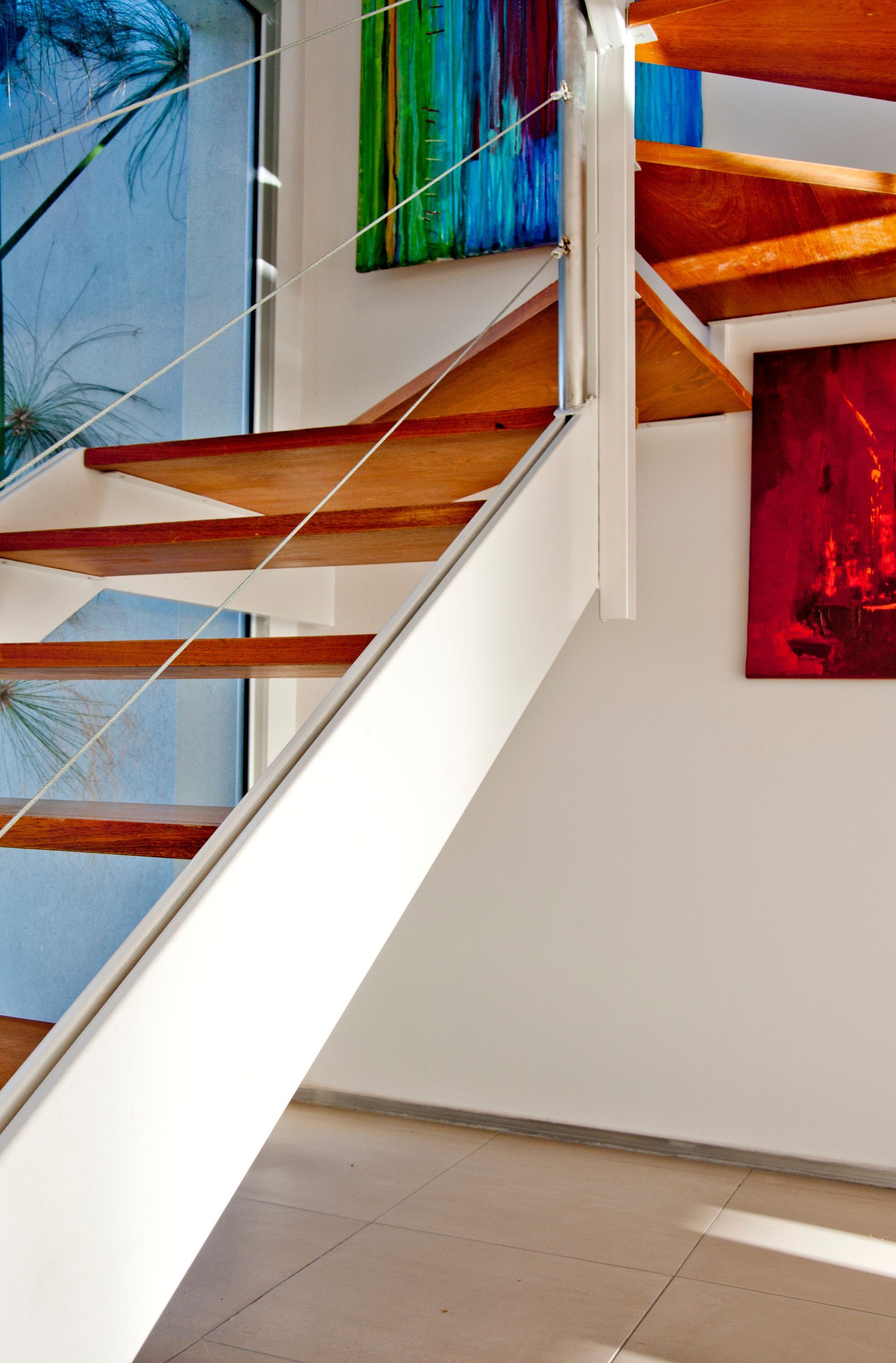 Escalera de madera con barandilla blanca y obras de arte coloridas sobre pared blanca.