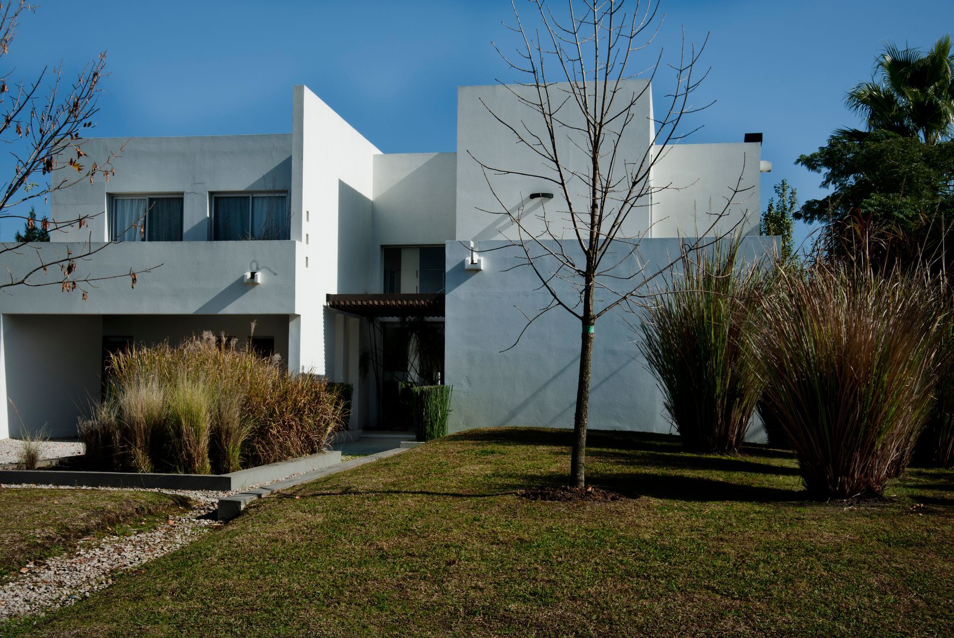 Casa blanca moderna con techo plano, pequeño patio, arbustos y un árbol desnudo contra un cielo azul.