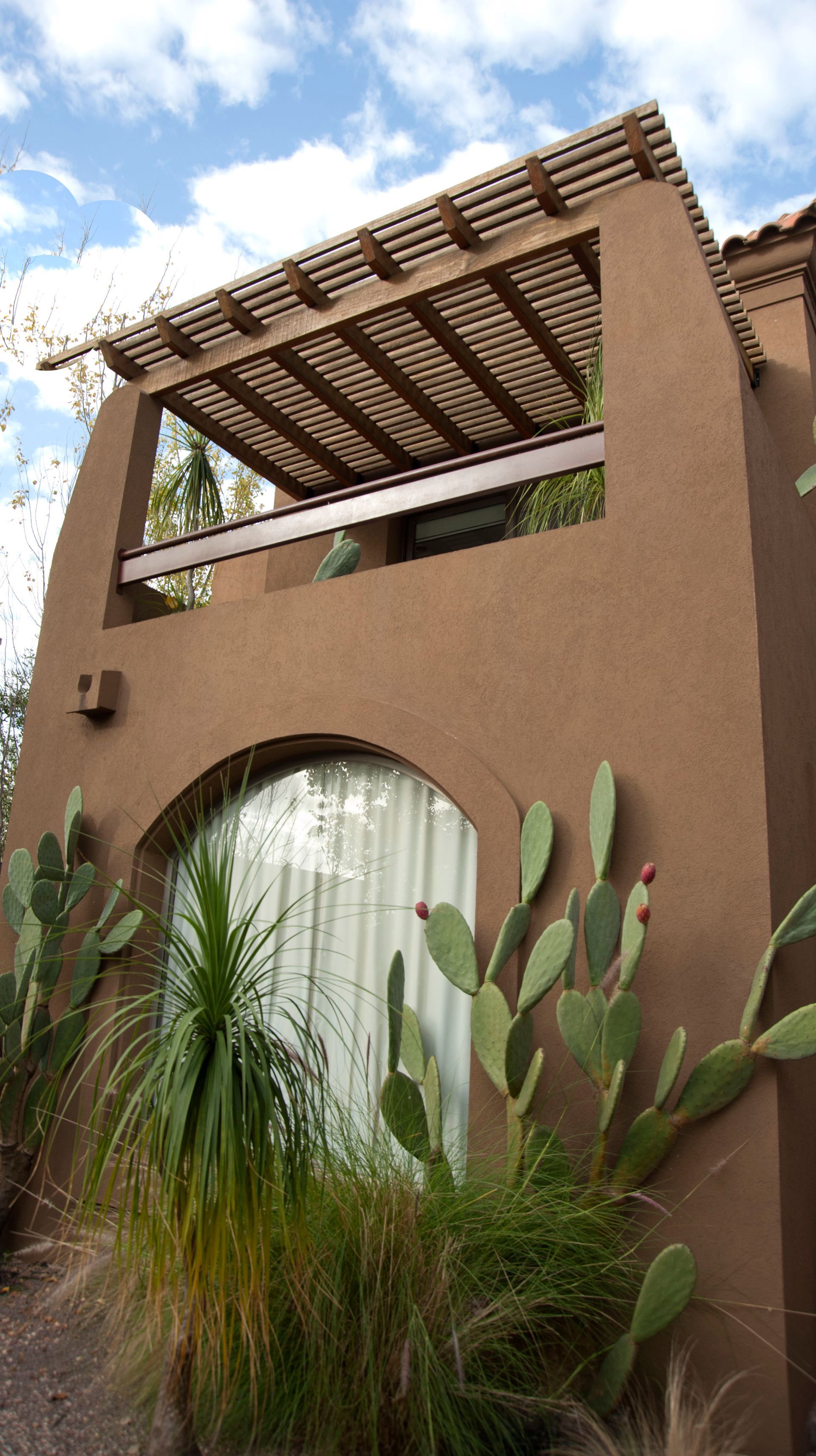 Edificio de adobe de dos pisos con balcón y pérgola de madera, cactus y plantas desérticas en primer plano.