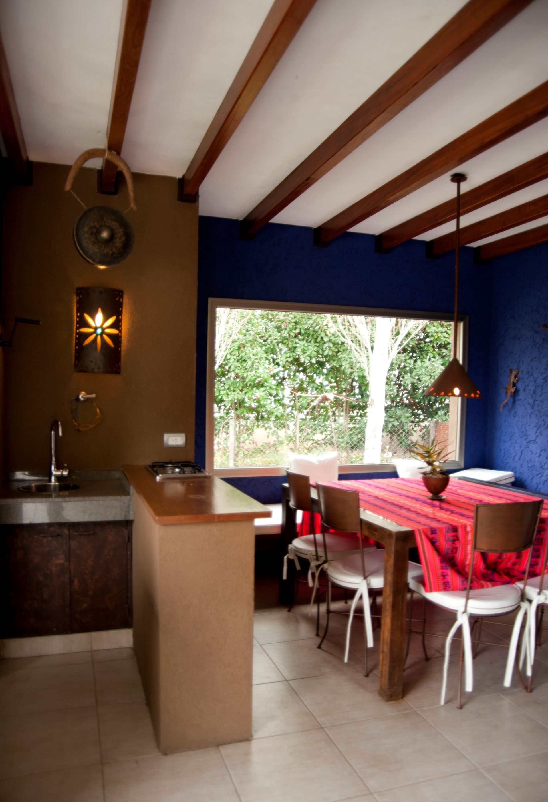 Cocina con comedor: paredes marrones y azules, vigas de madera, mesa puesta para comer, vista a la vegetación.