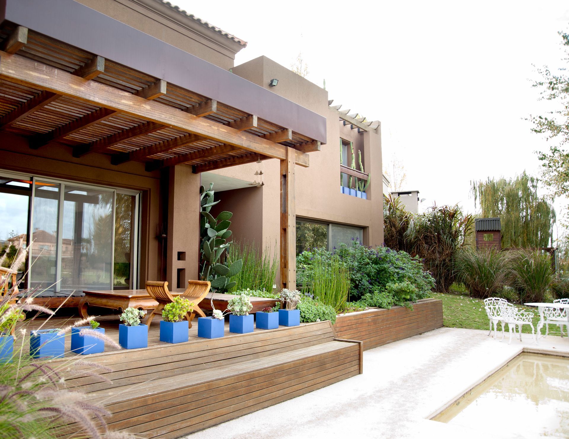 Exterior de casa con terraza de madera, jardineras azules y piscina.