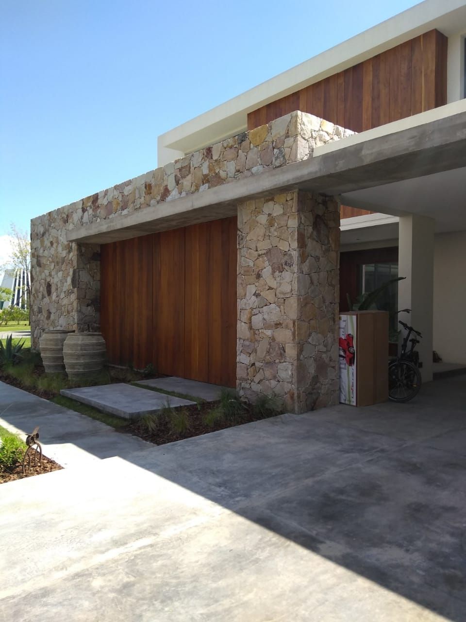 Exterior de casa moderna con fachada de piedra, puerta de garaje de madera, entrada de hormigón y un cielo brillante.