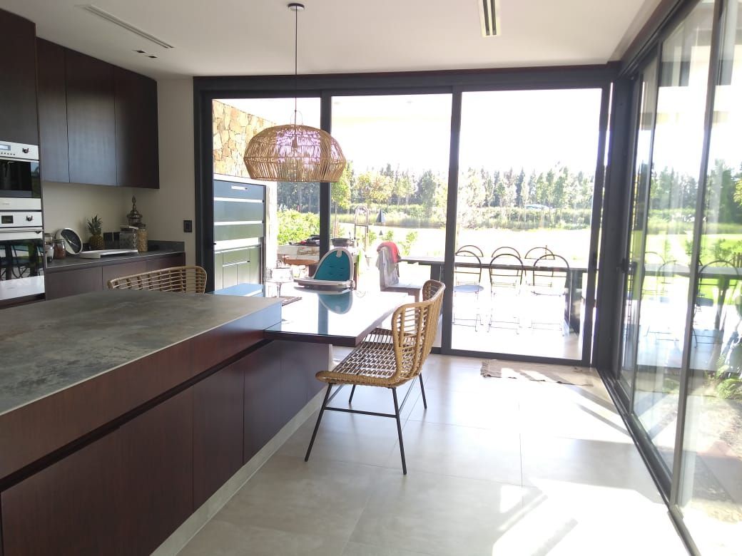 Cocina moderna con grandes ventanales, armarios oscuros y una silla de mimbre; a través de ellos brilla la luz del sol.