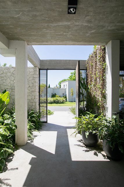 Entrada con exuberantes plantas, muro de piedra y vista a una calle soleada a través de un portón abierto.