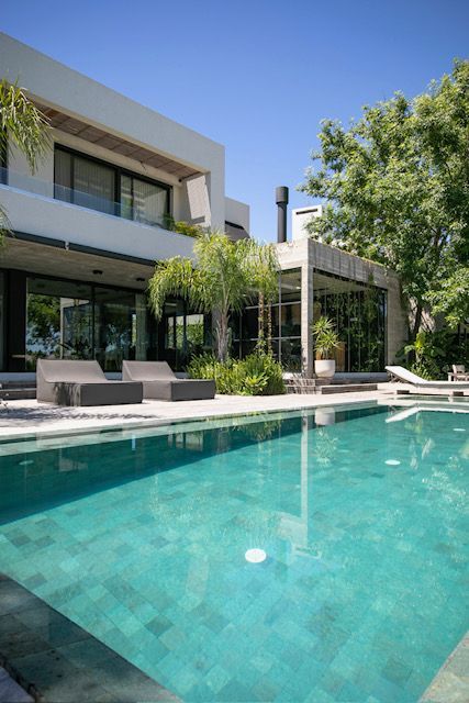 Casa moderna con piscina. Agua azul, paredes blancas, amplios ventanales, tumbonas y un soleado entorno exterior.