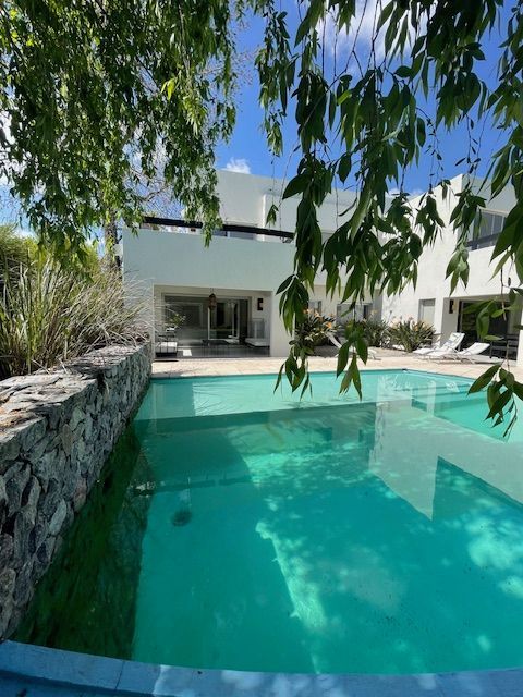 Piscina frente a un edificio blanco con un patio sombreado, cielo azul brillante visible.