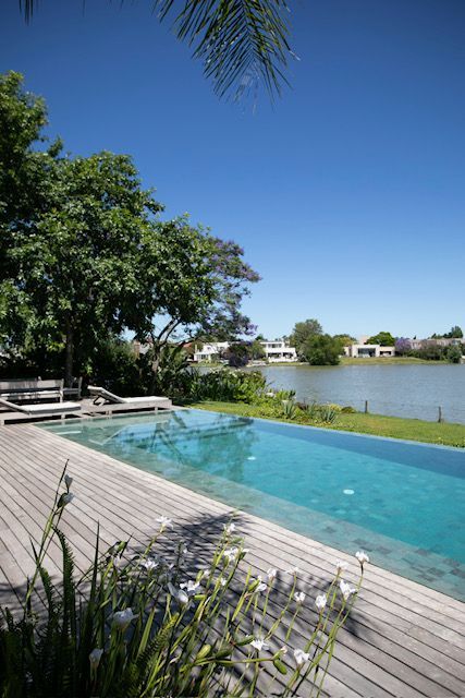 Vista de la piscina: piscina rectangular larga, terraza de madera, vegetación, lago, cielo azul claro.