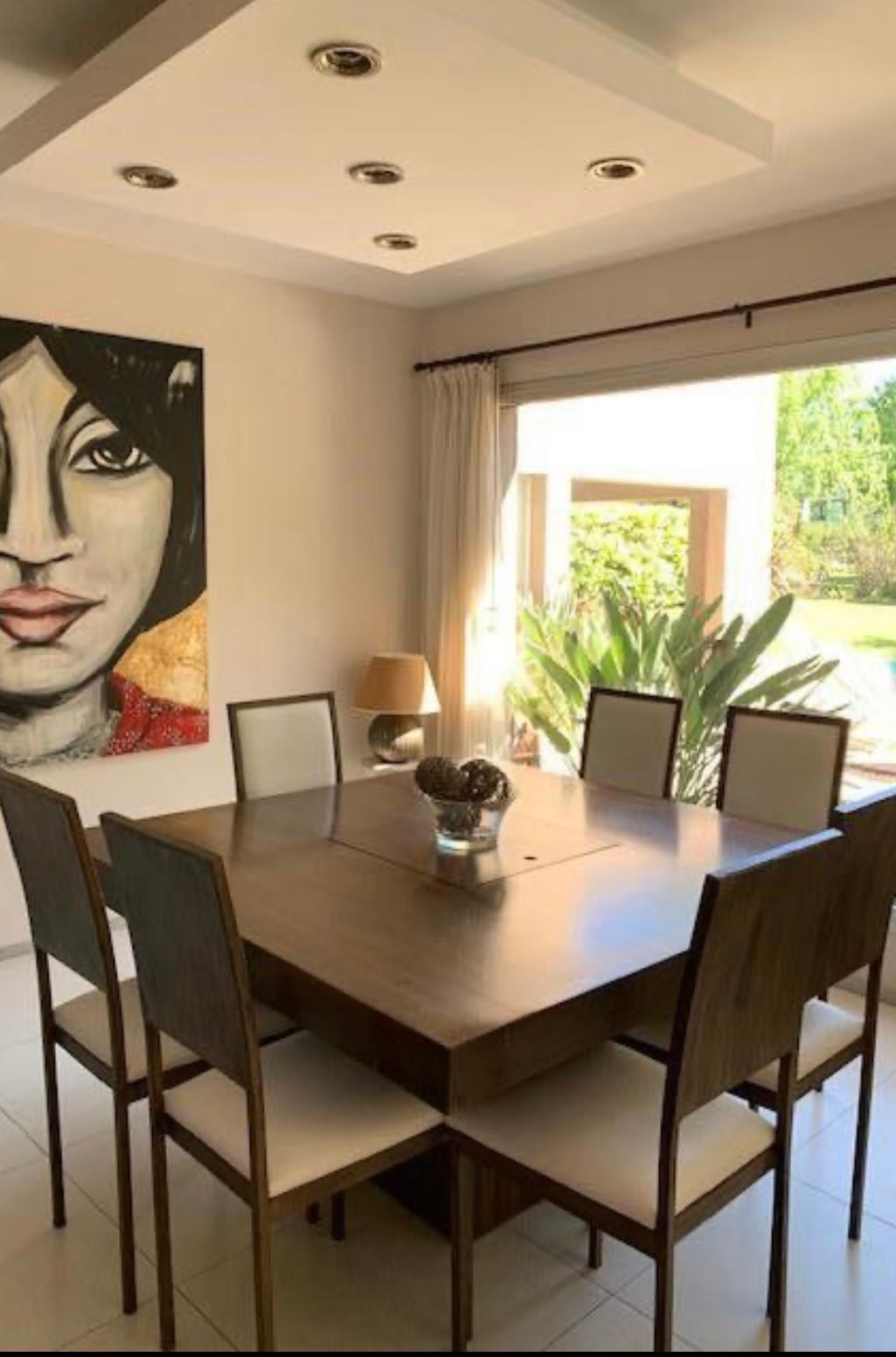 Comedor con una gran mesa de madera, ocho sillas y un cuadro abstracto; una ventana muestra una vista exterior.