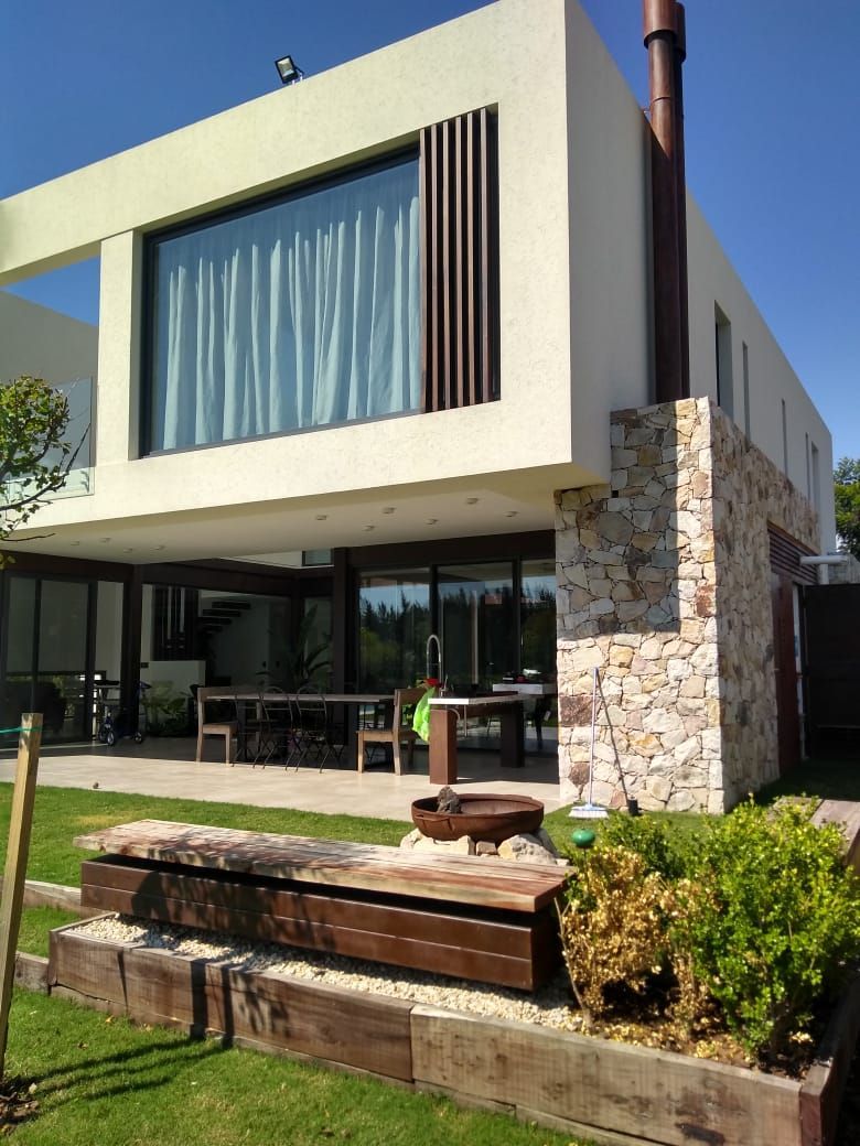 Casa moderna con un gran ventanal, detalles de madera y pared de piedra. Comedor exterior y jardín.