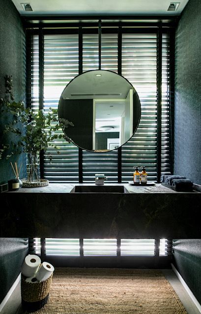 Baño en tonos oscuros con un gran espejo redondo, tocador oscuro y ventana con rejilla, con una cesta de papel higiénico.