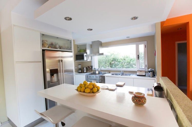 Cocina blanca moderna con electrodomésticos de acero inoxidable, barra de desayuno y una pared naranja.