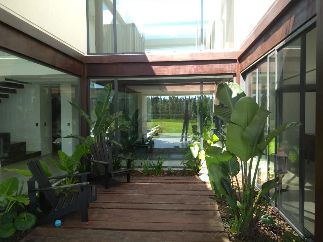 Pasarela de madera flanqueada por plantas y que conduce a una zona con paredes de cristal, muebles de exterior y césped.